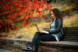 Kvinne sitter på en steinbenk med en laptop på fanget. Omgivelsene er høstlige. Til venstre for kvinnen og benken er en busk med røde blader. Gresset er gulnet.