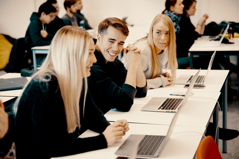 Foto av to kvinnelige og en mannlig student som sitter foran hver sin laptop på en skolepult i et klasserom.