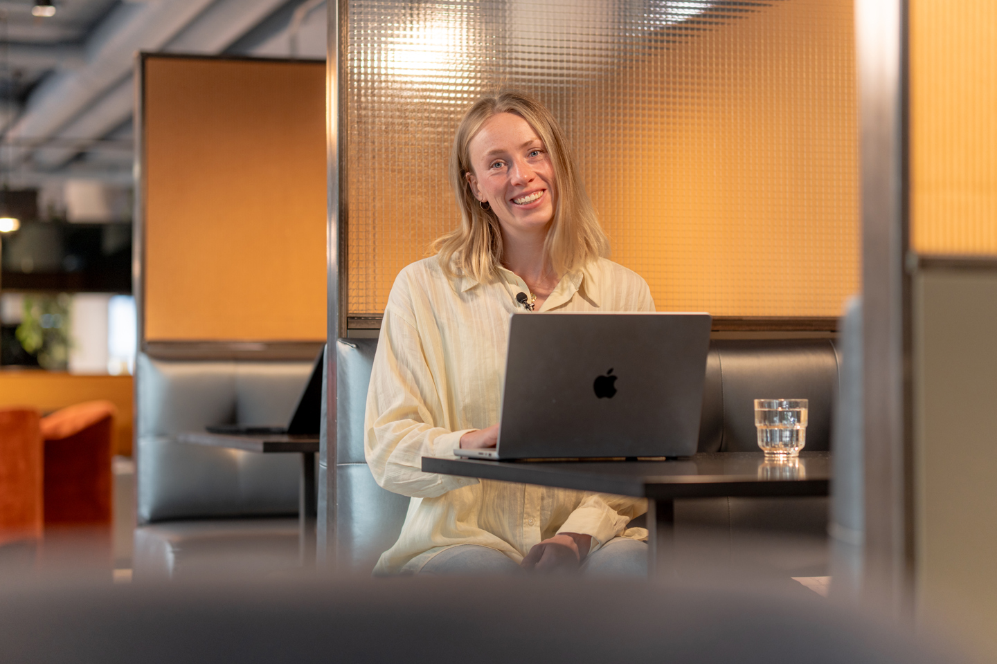 Person sitter på PC og smiler inn i kamera, i et kontorlandskap 