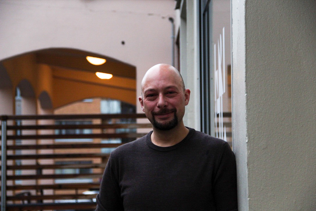 Professor Tor-Morten Grønli ser i kameraet og smiler. Han står med kroppen vendt mot kameraet og lener venstre arm mot en murvegg. Han har på seg en brun genser og er skallet. Murveggen er lys, i bakgrunnen ser man et gjerde og en undergang til en videre gate.