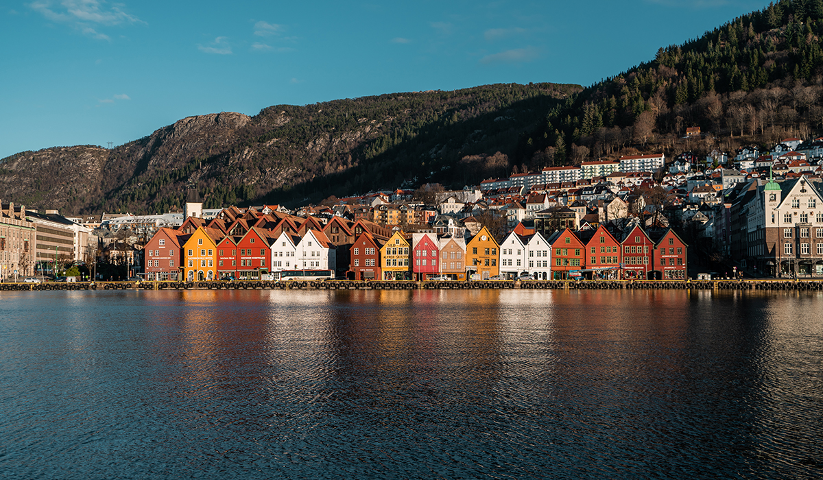 Bildet viser et bilde av Bryggen i Bergen. Solen stråler på de gamle husene og himmelen er blå. 