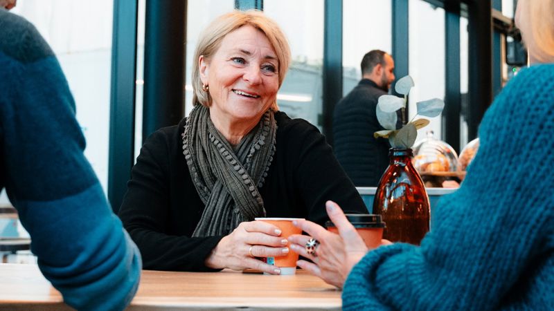 Kvinne sitter på kafe og snakker med to andre som sitter med ryggen til. 