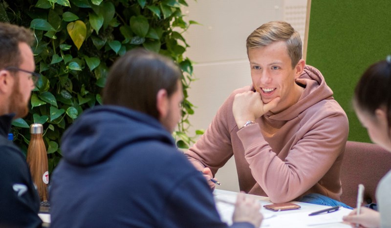 En gruppe studenter sitter rundt et bord. En mann i rosa hettegenser er i fokus. 