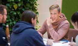 En gruppe studenter sitter rundt et bord. En mann i rosa hettegenser er i fokus.