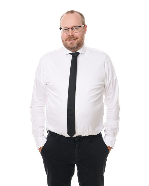 Mann med briller. hvit skjorte, svart slips, svarte bukser.