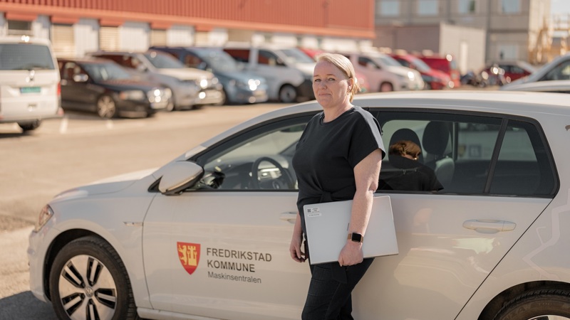 Person står med PC i hånda foran en hvit tjenestebil på en parkeringsplass.