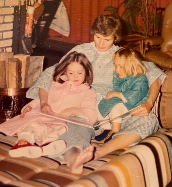 To små søstre sitter tett sammen med sin mamma når hun leser bok på tidlig 70-tall.