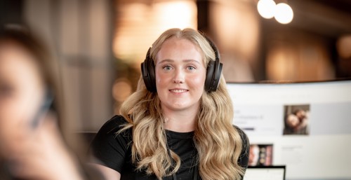 Person ser smilende inn i kamera med hodetelefoner i et kontorlokale