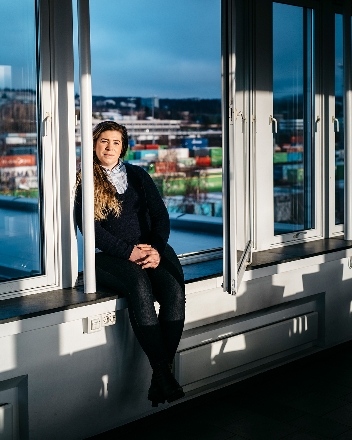 Bildet er tatt av Anna Svanberg som sitter i et vindu. Gjennom vinduet ser man sjø og containere på et konstruksjonsområde. Anna har på seg mørke bukser og mørk genser.