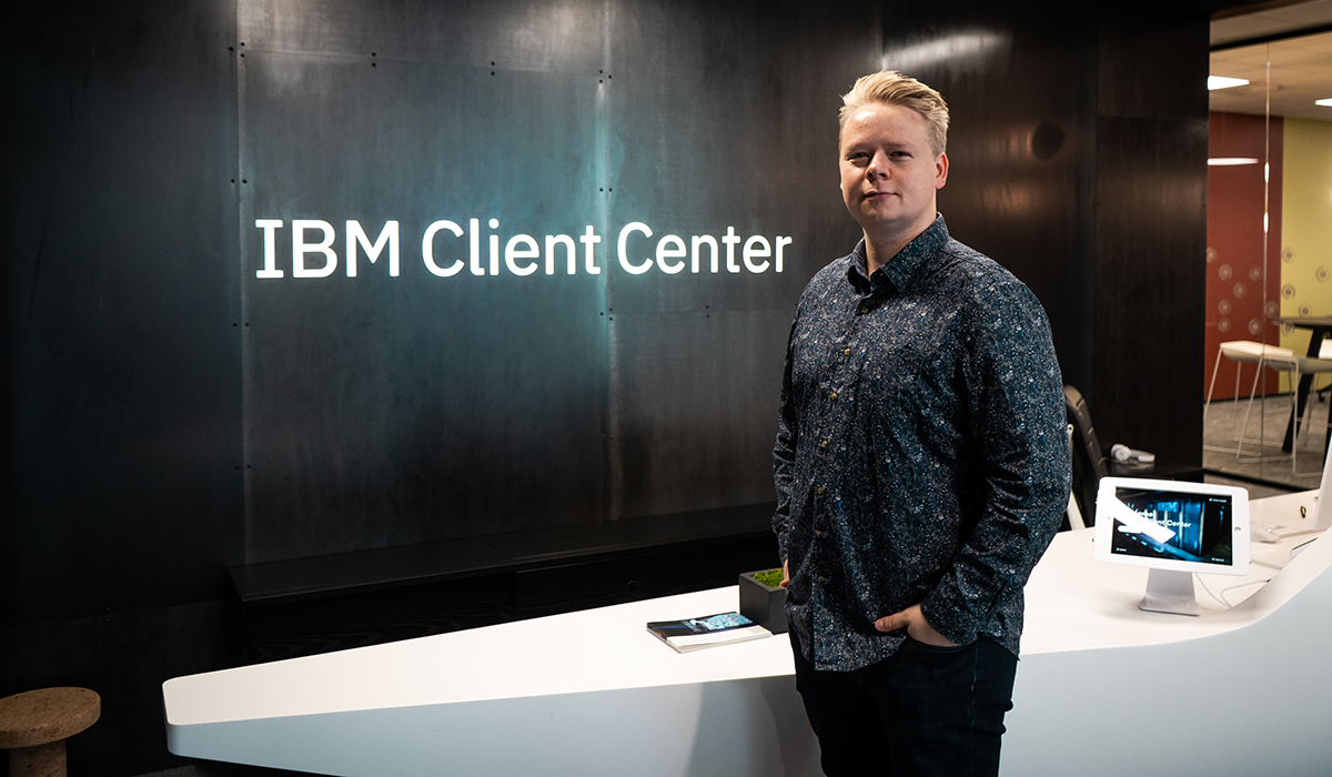 Jo Anders står foran skiltet til "IBM Client center" og en hvit resepsjonsbenk står bak han og veggen bak er mørk. Jo Anders har hendene i lomma og har på seg en mørkeblå skjorte og sorte jeans.