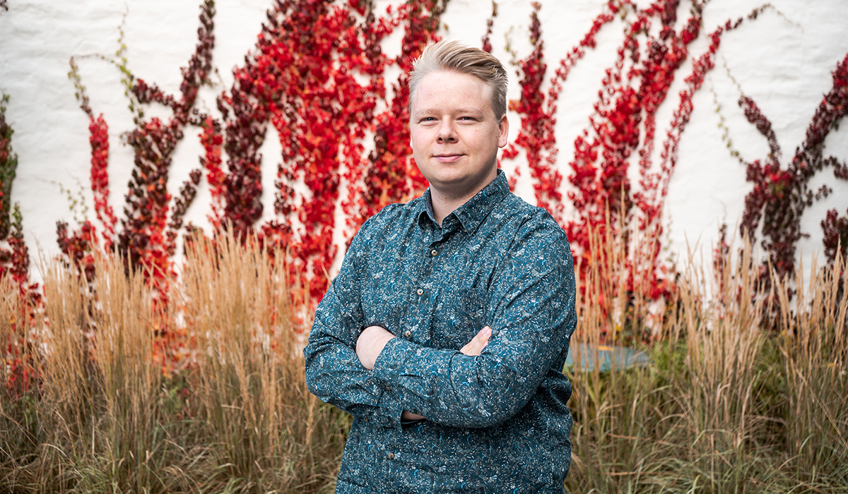 Jo Anders står utendørs med armene i kryss over brystet. Han har på seg en mørkeblå skjorte og har lys hud. Bak han er det knallrøde høstblomster som står opp av bakken. Det er også brunt høyt gress bak han.