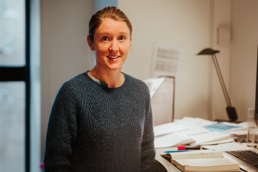 Foto av kvinne i blå genser oppstilt ved siden av en kontorpult.