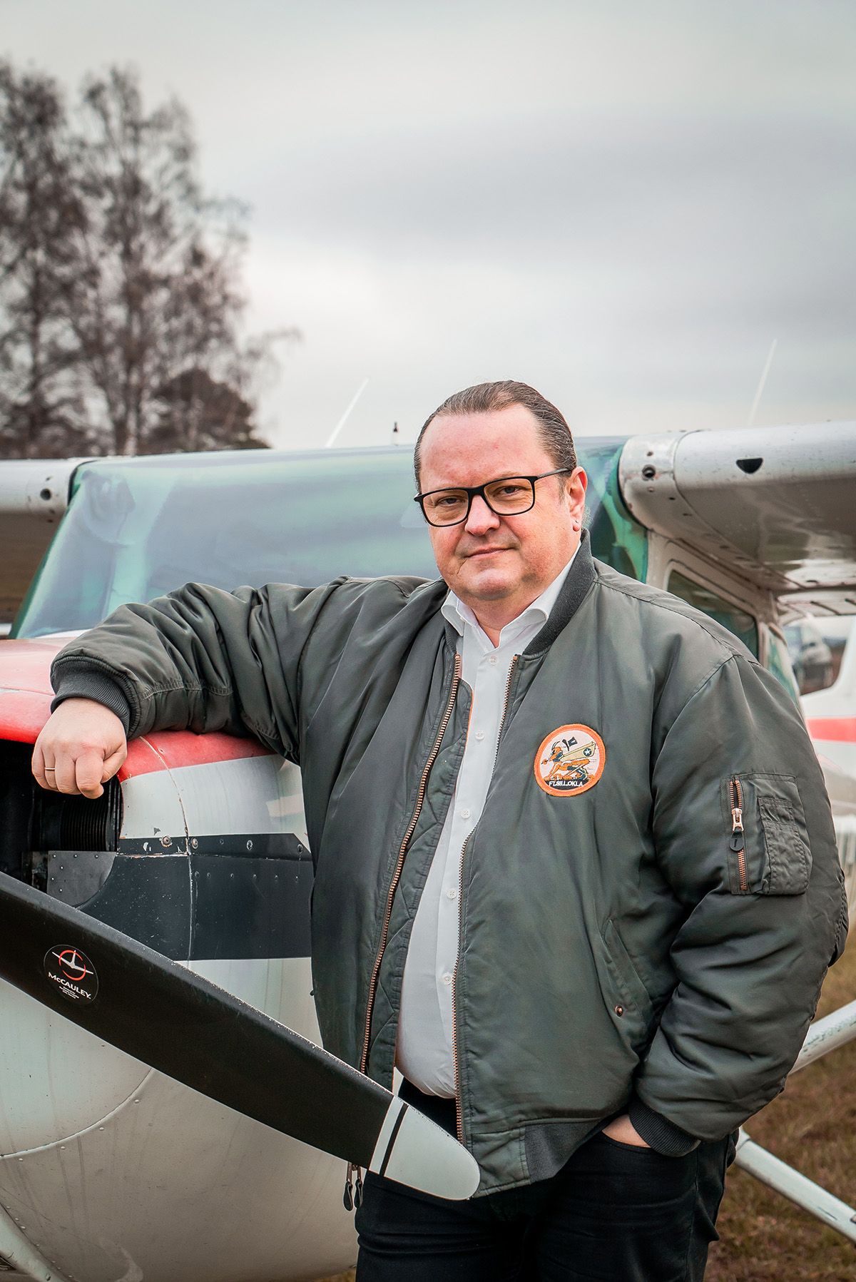 Arne H. Krumsvik står foran et småfly på Kjeller flyplass utenfor Oslo. Han har på seg en mørkegrønn bomberjakke. Han har den ene armen over snuten på miniflyet. Bildet viser et nærbilde av han. 