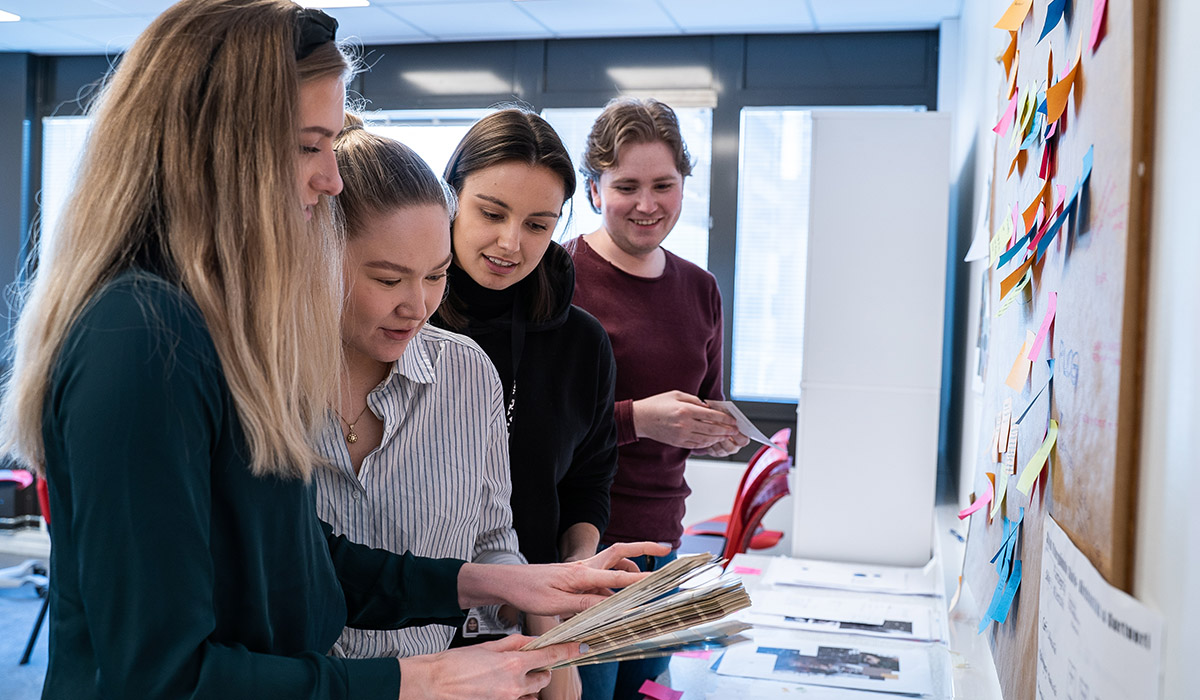 Foto av fire interiørstudenter ved Fagskolen Kristiania, som ser på materialprøver. Foran dem henger en tavle med mange fargerike post it-lapper.  
