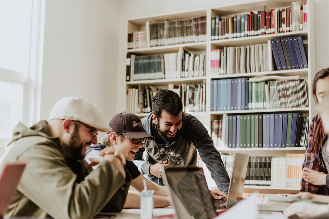 Three men smiling by computer surrounded by books on shelf