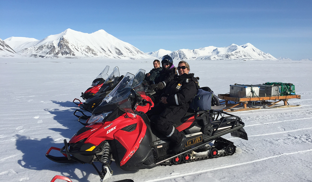 Elin Bolann og kollegaene på snøscootere i Svalbard i forbindelse med forskningsprosjektet i Adventure Tourism. Scøscooterene er røde, tre stykker, med blå himmel og hvite fjell bak. Bakken er snøbelagt. Alle tre menneskene ser inn i kameraet og smiler.
