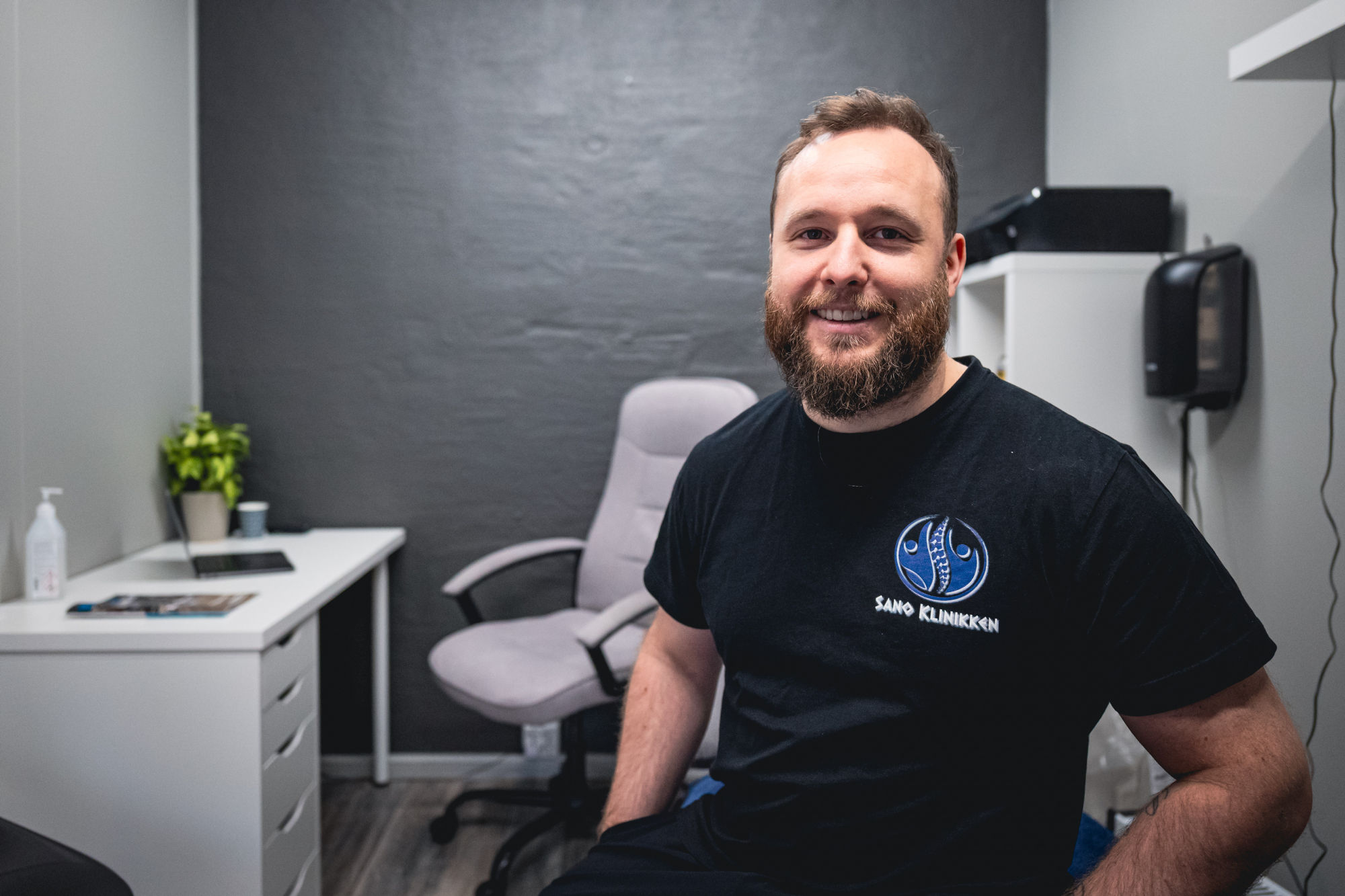 Marcus sitter på kontoret sitt i klinikken. Han har svart t-skjorte med klinikkens logo.