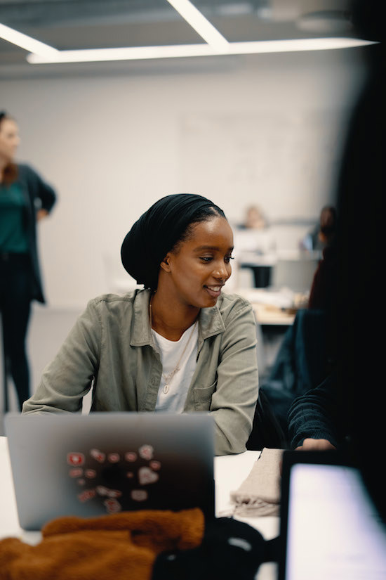 Student sitter foran laptop i klasserom og ser på en medstudent. Personen smiler og følger med på hva studenten har å vise.