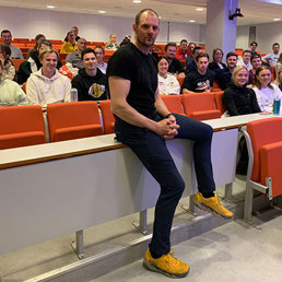 Mann hviler seg oppå en pult, bak sitter studenter på rekke i et auditorium.