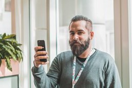 Mann med skjegg står og ser inn i kamera med en telefon i hånden. 