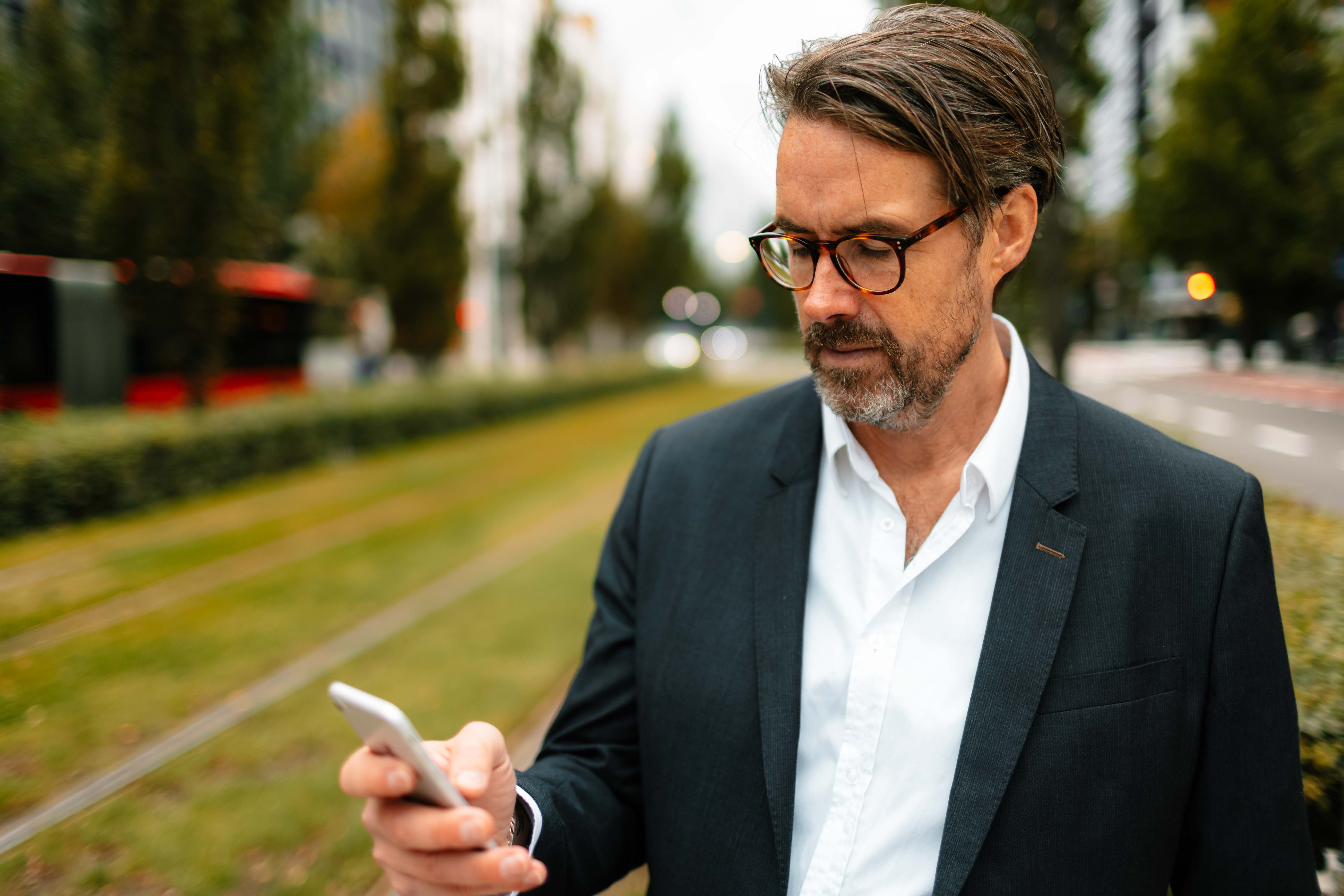 En middelaldrende mann med briller, svart dressjakke og hvit skjorte står foran en trikkeskinne og sjekker mobilen.