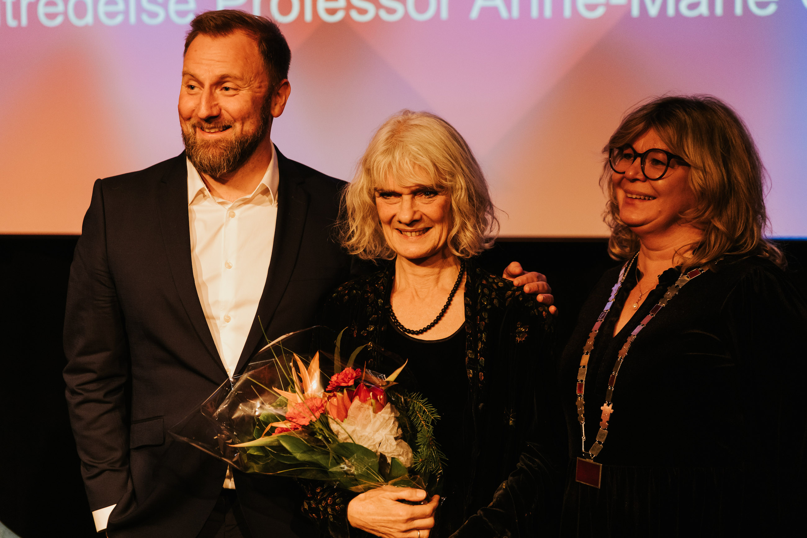 Anne-Marie Giørtz står i midten og smiler med blomster. På venstre side står em blid Kai Hanno Schmidt og på høyre side står prorektor Trine Johansen Meza og ser glad ut.
