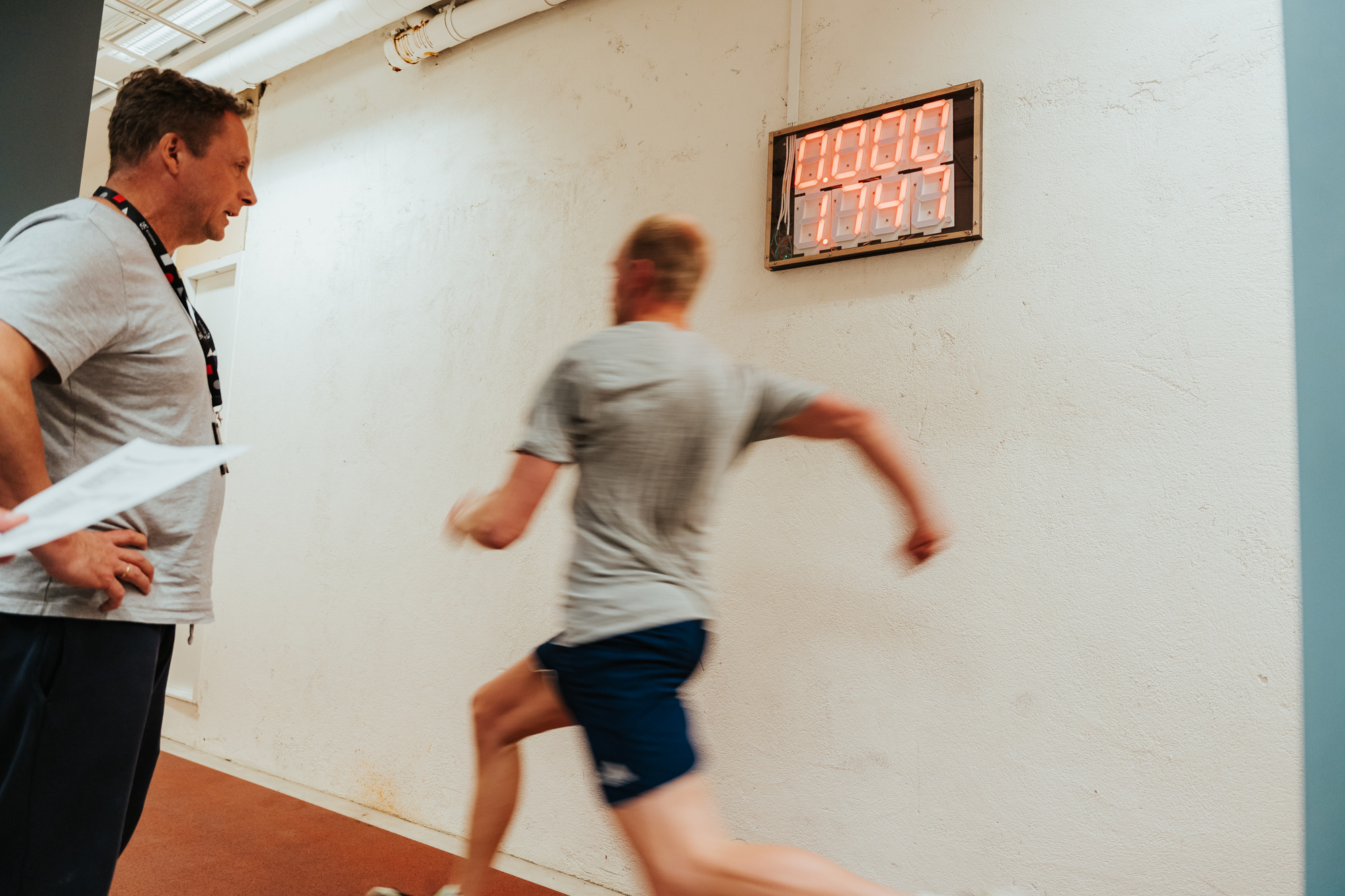 En trener iført sportsklær står med ryggen til kamera og observerer en mann som løper på en innendørs løpebane. 