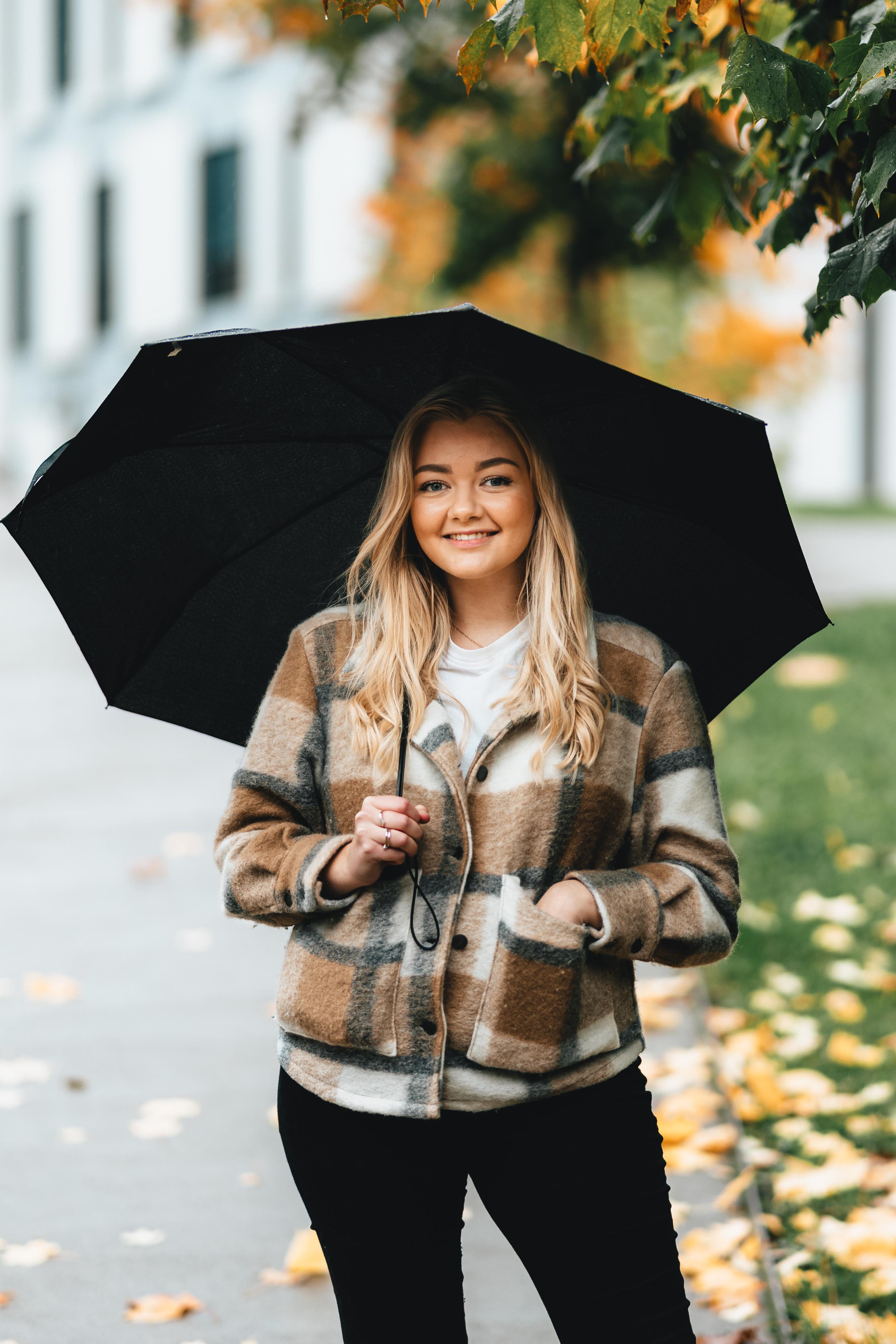 Bilde av ung blond kvinne med rutete høstjakke og paraply som står under et tre i regnvær.