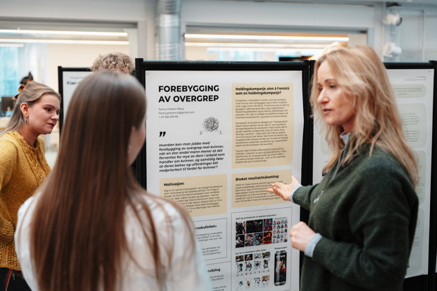 Mennesker diskuterer foran designarbeider hengt opp på en skillevegg.
