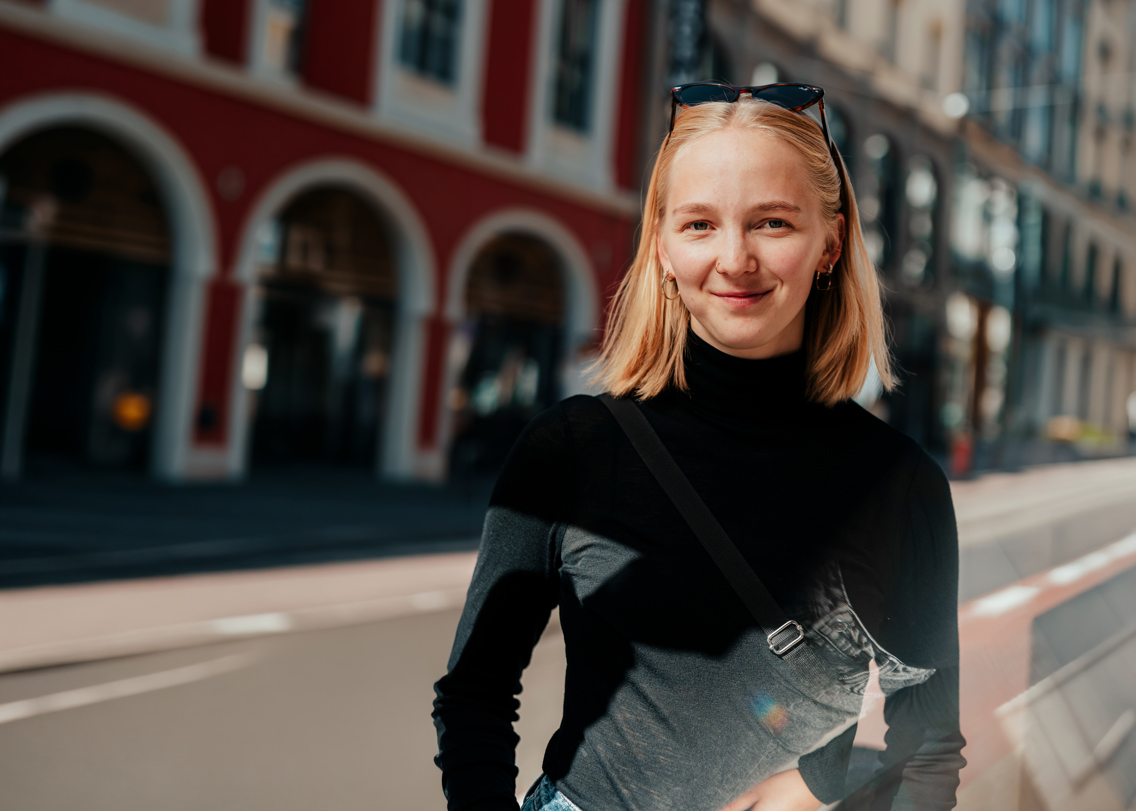 Kvinne i svart tøy, med solbriller på hodet, står og smiler til kamera i en tom gate