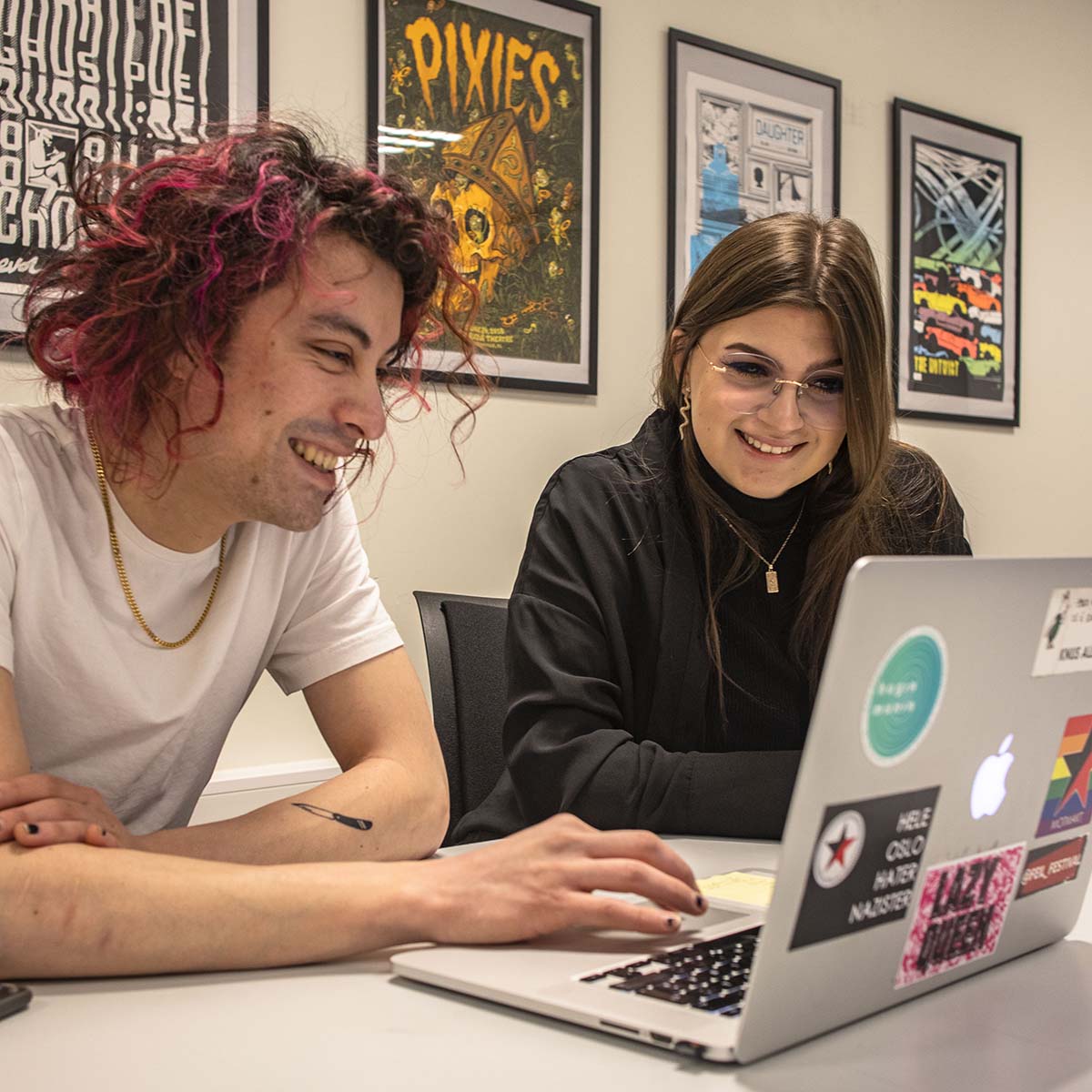 To studenter sitter og smiler til en laptop. Han har rosa hår og hun har mørkt hår og briller. 