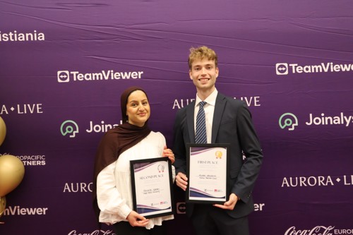 To studenter står foran et banner med hvert sitt diplom