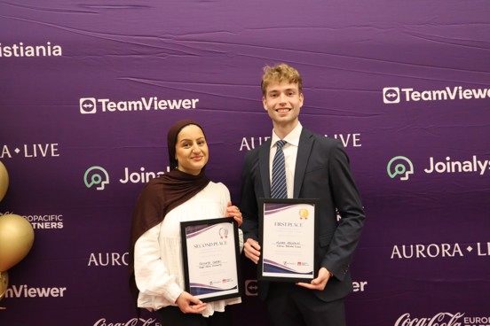 To studenter står foran et banner med hvert sitt diplom