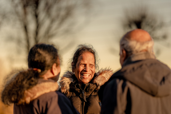 Tre personer står utendørs i varme jakker og snakker sammen i et gyllent kveldslys.