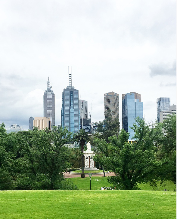 Bildet viser en linje med skyskrapere i Melbourne. Foran skyskraperne kan man se grønt gress og en park med tær.