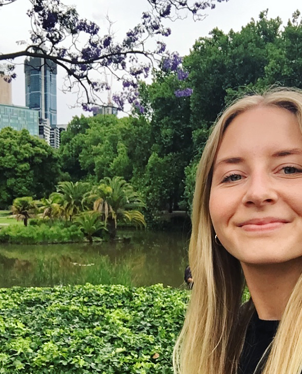 Matilde står i en park i Melbourne. Bak henne kan man se en liten innsjø og frodige grønne planter og trær. Mathilde har lagt lyst hår og har på seg en sort t-skjorte.