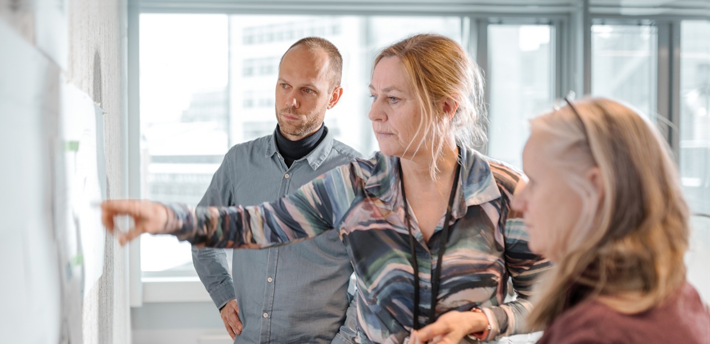 Tre personer ser på en prosjekttavle i et lyst kontorlokale