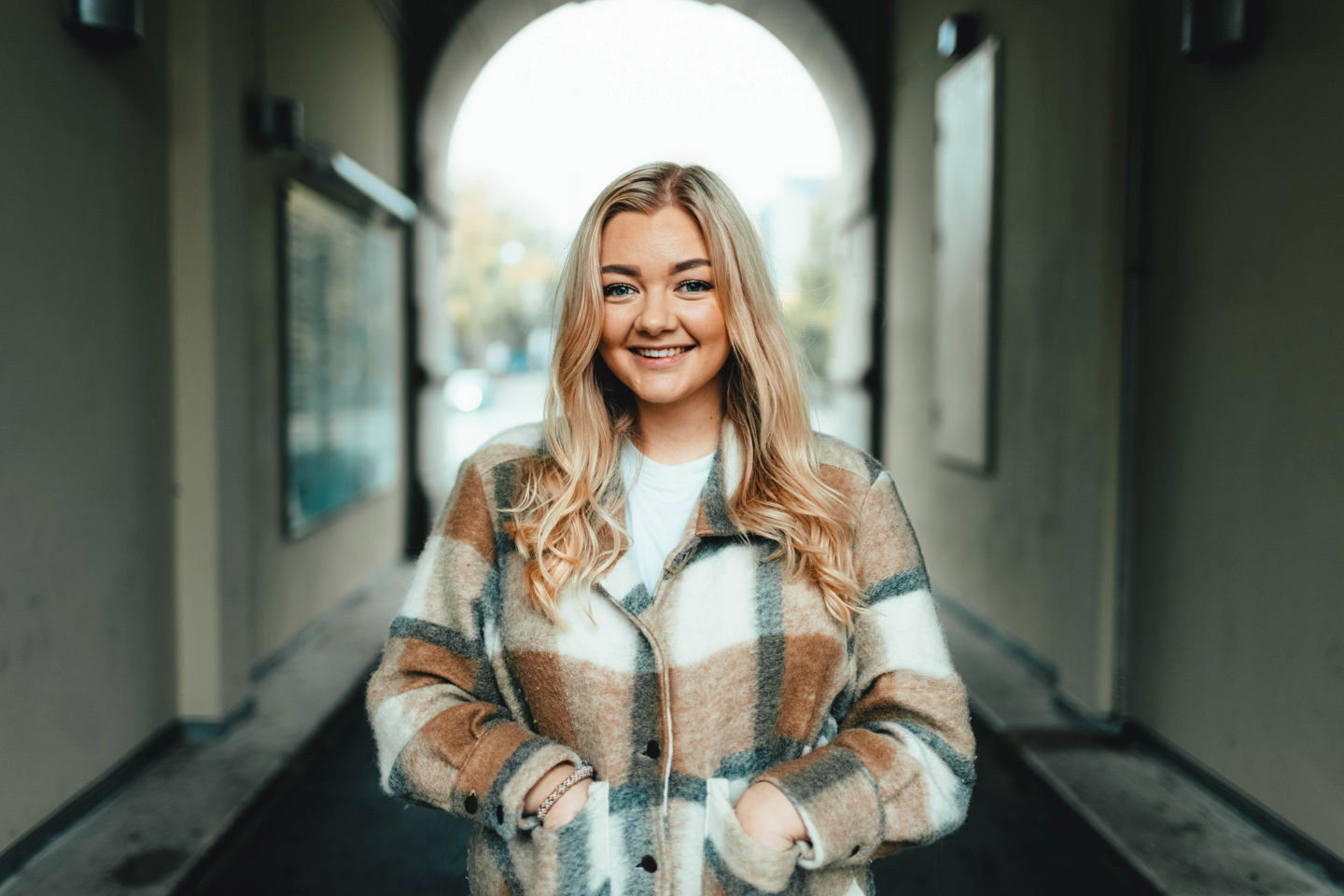 Bilde av ung blond kvinne i rutemønstret høstjakke som står i en passasje.