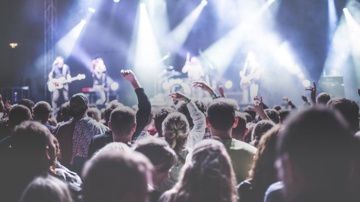  Bildet viser et publikum ved en konsert, vendt mot en scene med lysglimt og uskarpe musikere. En person i mengden løfter en arm, et tegn på feiring og glede. Atmosfæren er festlig og energisk.