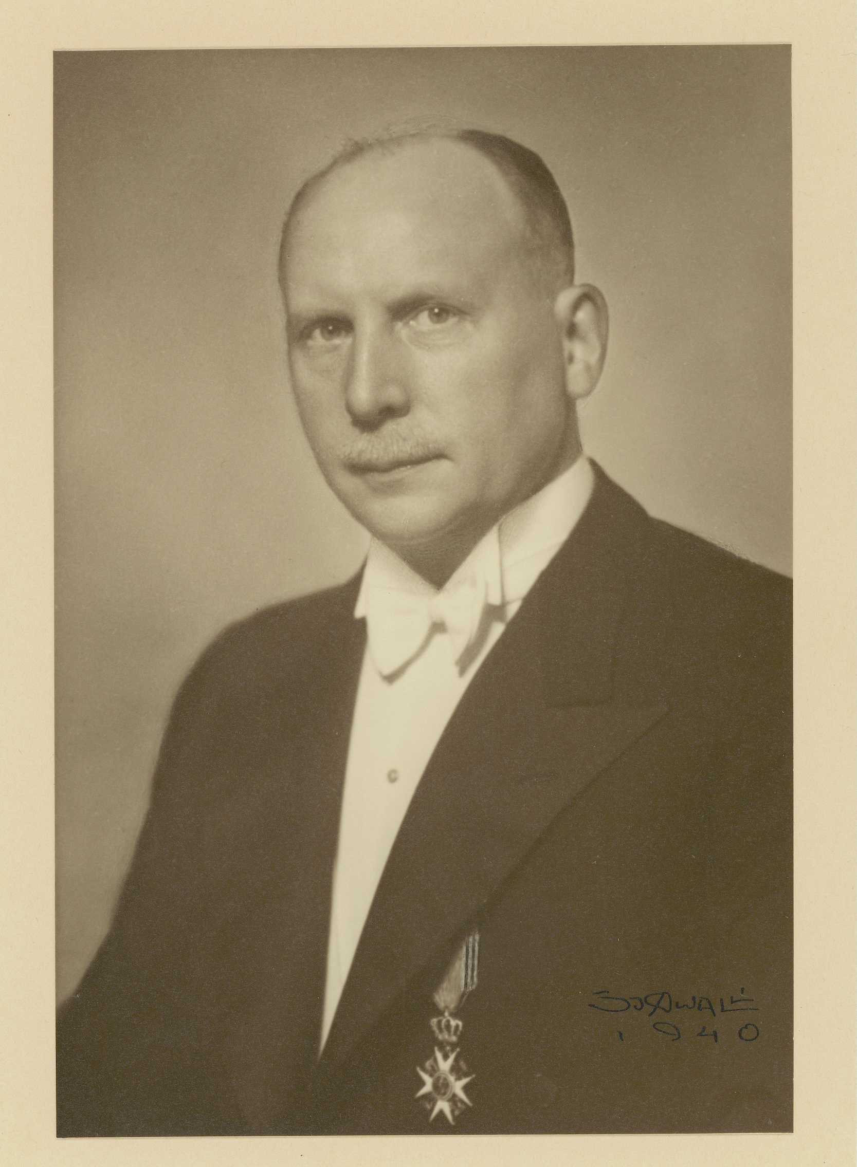 En høytidelig mann sitter i dress med medaljen St. Olavskorset på jakkeslaget.