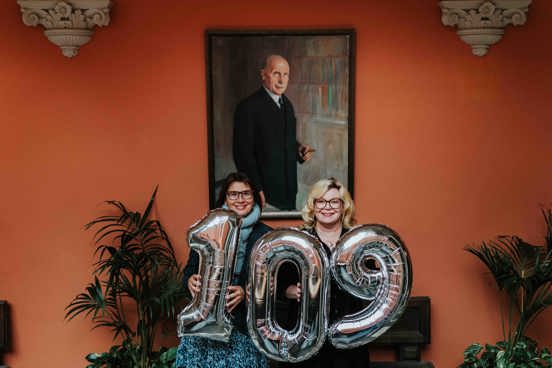 Administrerende direktør Solfrid Lind og rektor Trine Johansen Meza står foran portrettet av Ernst G. Mortensen, med oppblåste tall i hendene som til sammen viser 109. 