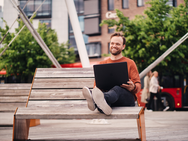 Mannlig nettstudent sitter ute med laptopen i fanget. I bakgrunnen kan man se folk som går på en buss.