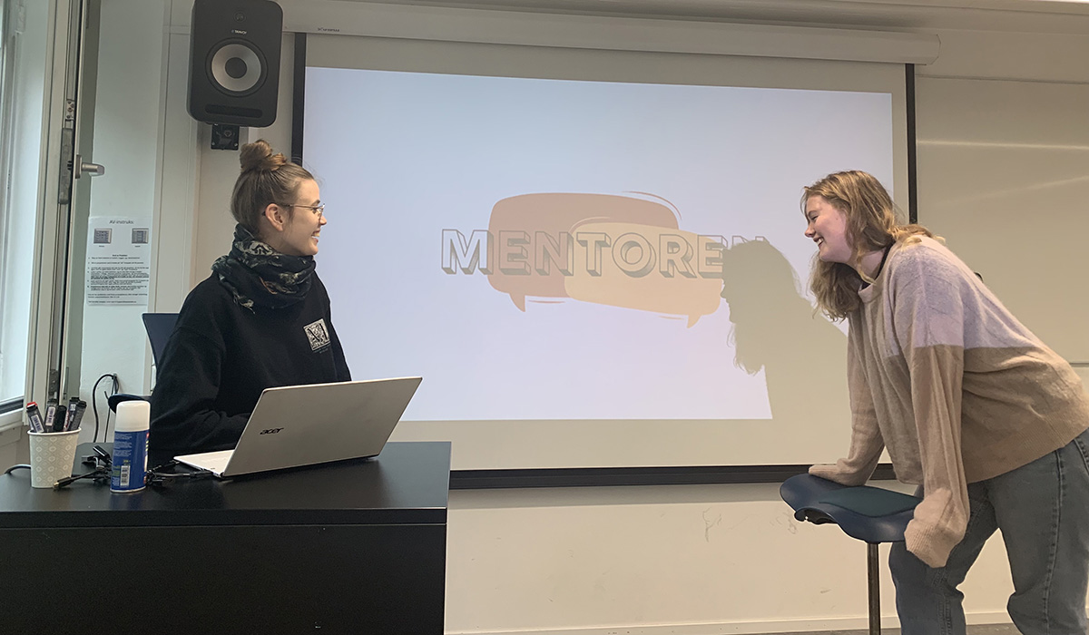 Ida Kristine Jorddal og Nora Strønstad Elder står i et klasserom. I bakgrunnen ser man presentasjonen av podcast-konseptet Mentoren.