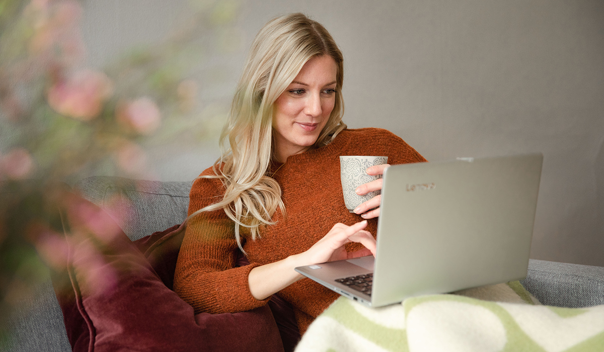 Kvinne sitter i sofaen hjemme med kaffekopp, laptop i fanget og studerer på nett