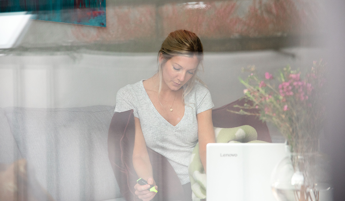 Kvinne sitter i sofaen hjemme med laptop i fanget og studerer på nett