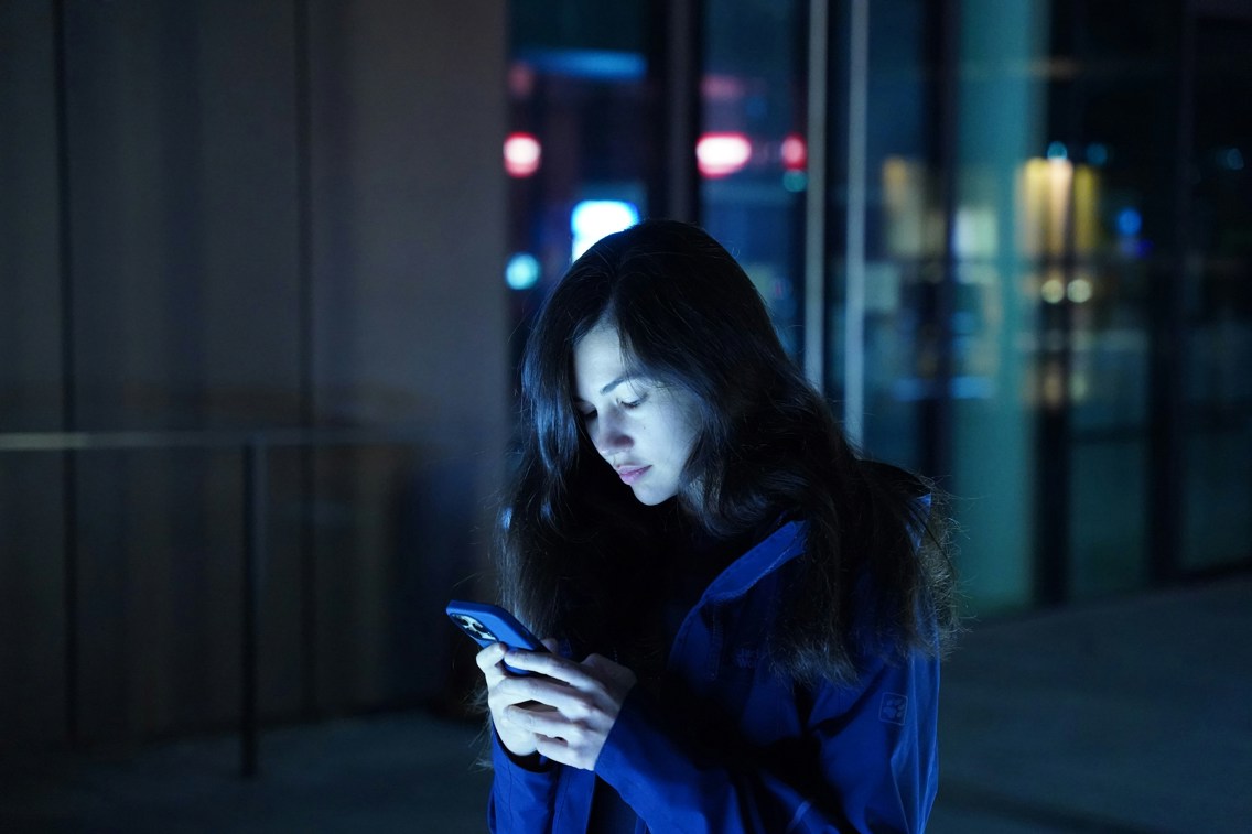 Kvinne som står ute i mørket og ser ned på telefonen sin