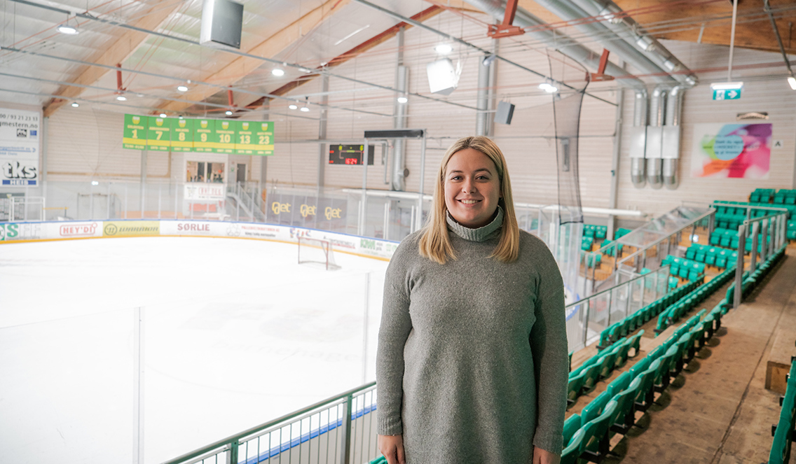 Cecilie Olsen står på en tribune. Bak henne kan man se en ishockey-bane og stoler. 