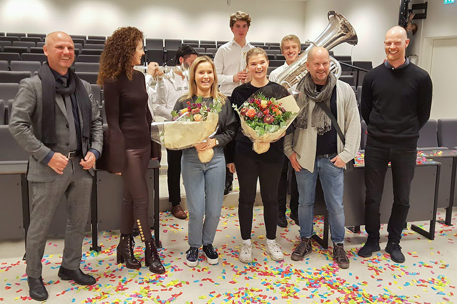 Interiørstudentene ved Fagskolen Kristiania, Eilén Benedikte Brenne og Hanne Lassemo Buøen står side om side med juryen til Clarion Hotel The Hub. Studentene holder hver sin blomsterbukett. I bakgrunnen står et musikkorps, og på gulvet ligger det konfetti. 