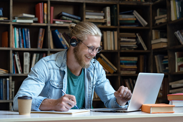 Mann med headset i et bibliotek som jobber på PC og noterer i en bok
