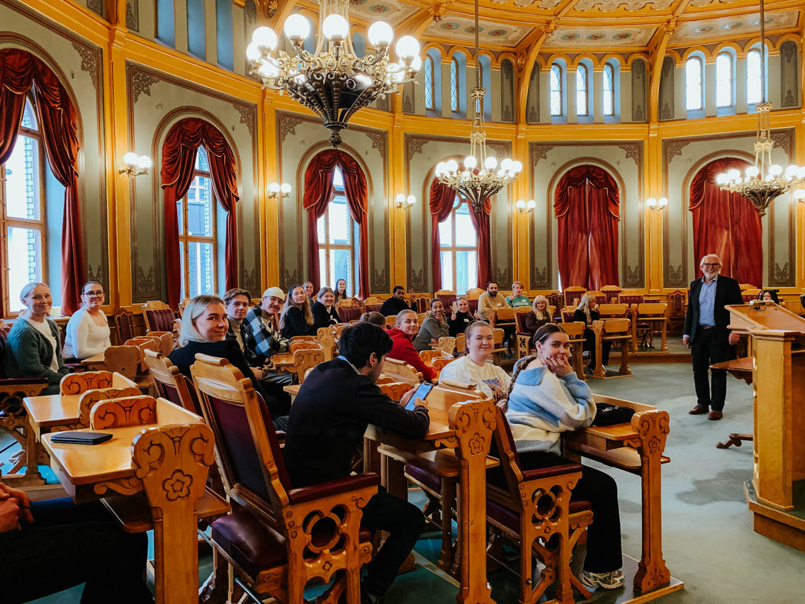 En gruppe studenter er plassert i stortingssalen sammen med Carl Erik Grimstad. De smiler mot kamera. Foto.